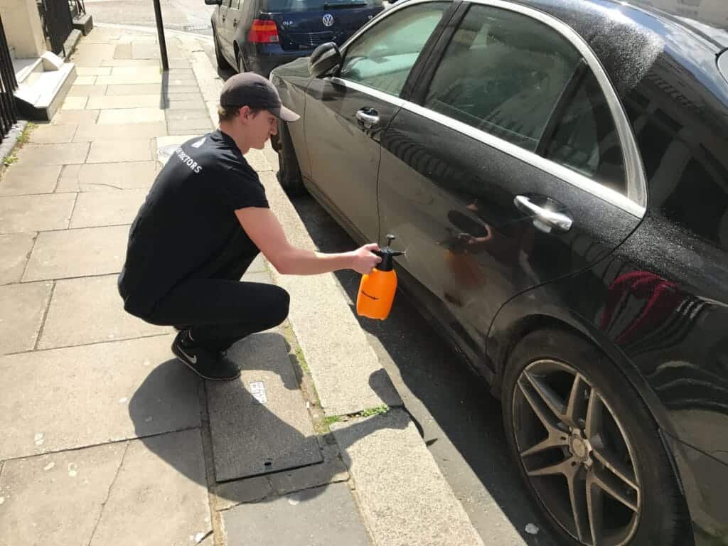 Technician cleaning a black car with spray bottle on a city street.